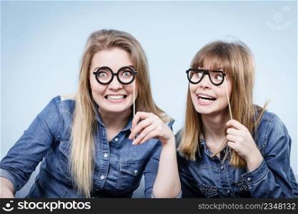 Two happy women holding fake eyeglasses on stick having fun fooling around wearing jeans shirts. Photo and carnival funny accessories concept.. Two happy women holding fake eyeglasses on stick