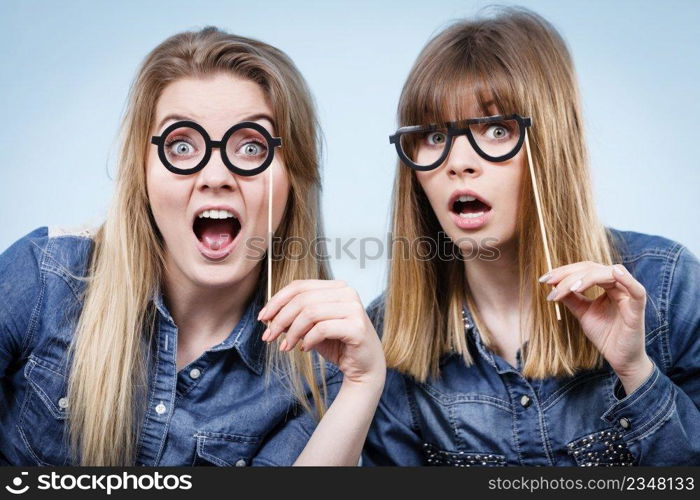 Two happy women holding fake eyeglasses on stick having fun fooling around wearing jeans shirts. Photo and carnival funny accessories concept.. Two happy women holding fake eyeglasses on stick