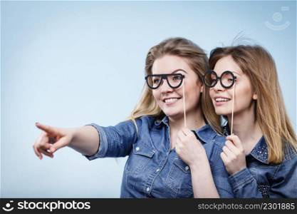 Two happy women holding fake eyeglasses on stick having fun pointing somewhere wearing jeans shirts. Photo and carnival funny accessories concept.. Two happy women holding fake eyeglasses on stick