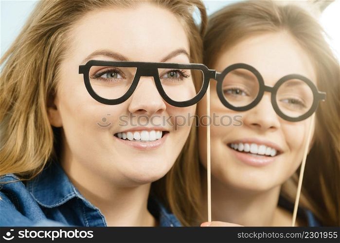 Two happy women holding fake eyeglasses on stick having fun wearing jeans shirts. Photo and carnival funny accessories concept.. Two happy women holding fake eyeglasses on stick
