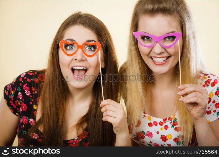 Two happy women holding fake eyeglasses on stick having fun wearing tshirts with flower pattern. Photo and carnival funny accessories concept.. Two happy women holding fake eyeglasses on stick