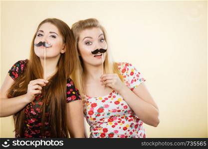 Two happy women holding fake moustache on stick having fun wearing tshirts with flower pattern. Photo and carnival funny accessories concept.. Two happy women holding fake moustache on stick