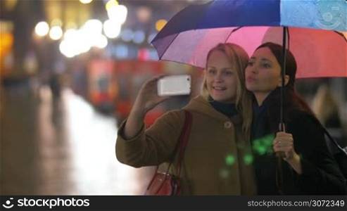 Two happy women making phone selfie under colorful umbrella during their friendly evening walk in the rainy city