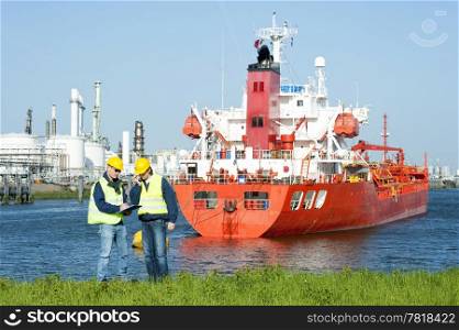 Two harbor workers at a petrochemical refinary harbor hard hats and safety vests discussing docking schedules