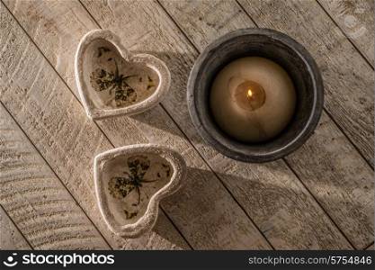 Two heart decorations on a white whashed wooden table next to a lit candle in a stoneware pot all in low directional light of the late afternoon.