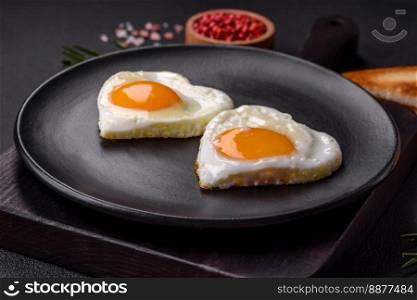 Two heart-shaped fried eggs on a black ceramic plate on a dark concrete background. Breakfast for valentine’s day
