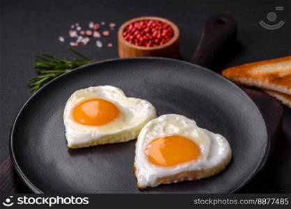 Two heart-shaped fried eggs on a black ceramic plate on a dark concrete background. Breakfast for valentine&rsquo;s day
