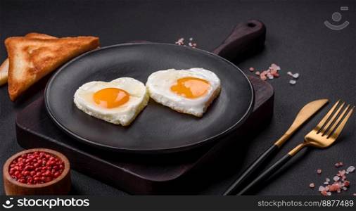 Two heart-shaped fried eggs on a black ceramic plate on a dark concrete background. Breakfast for valentine&rsquo;s day