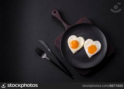 Two heart-shaped fried eggs on a black ceramic plate on a dark concrete background. Breakfast for valentine’s day