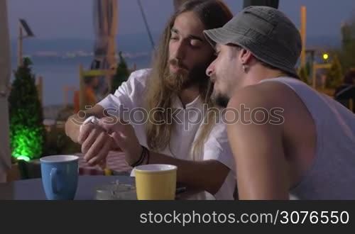 Two hippie man are speaking about white smart watch on hand of one and showing its functions against outdoor background sea sunset in cafe Thessaloniki, Greece