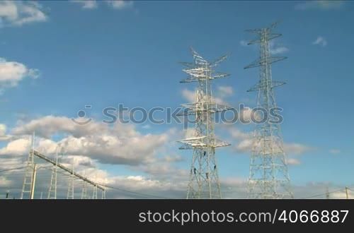 Two huge electrical towers with workers and clouds going by. Four workers are assembling the towers at the beginning of the power line. Electrical towers are an icon of our time. Suitable for development, technification, evolution, progress, future ideas, civilization, technological advance, town planning, modernization, human power, construction, economic expansion, prosperity, wealth, wealthy.