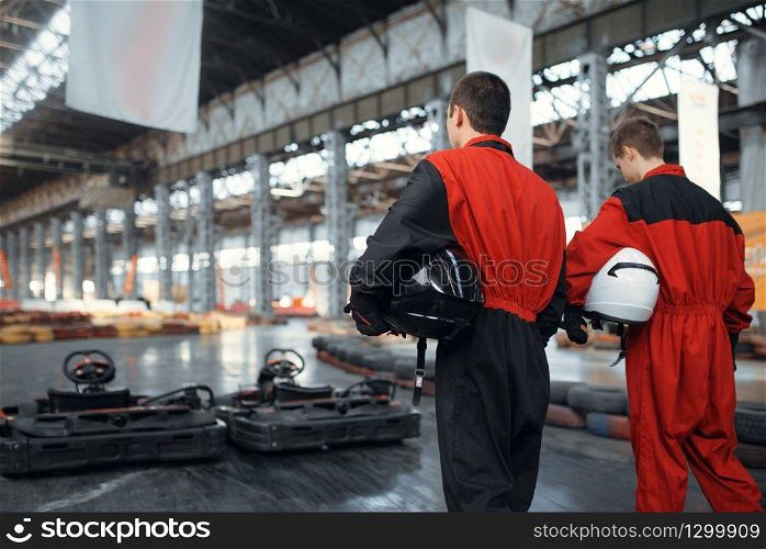 Two kart racers holds hemets, back view, karting auto sport indoor. Speed race on close go-kart track with tire barrier. Fast vehicle competition, high adrenaline leisure