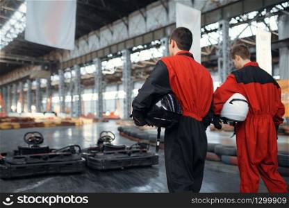 Two kart racers holds hemets, back view, karting auto sport indoor. Speed race on close go-kart track with tire barrier. Fast vehicle competition, high adrenaline leisure