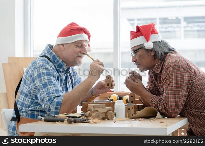Two kind senior carpenter men Caucasian and asian with mustache and beard wearing santa hat painting color with brushes on wooden toy house together at workshop on Christmas holiday