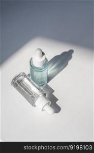 Two laconic bottles of cosmetic liquid in the rays of the harsh sun. A transparent white bottle with a pipette and a transparent blue one.. Two laconic bottles of cosmetic liquid in the rays of the harsh sun.