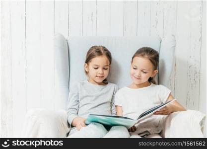 Two little adorable sisters sit in sofa, read interesting book, sit in comfortable sofa, enjoy togetherness. Best friends female children hold book, view pictures and read hearders, have spare time