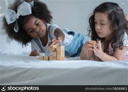 Two little diverse kid girls, African and Asian children enjoy playing build wooden block together on bed at bedroom