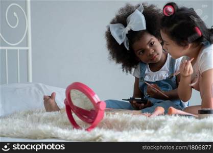 Two little girls, African and Asian children enjoy playing mother?s cosmetic make up together on bed at bedroom