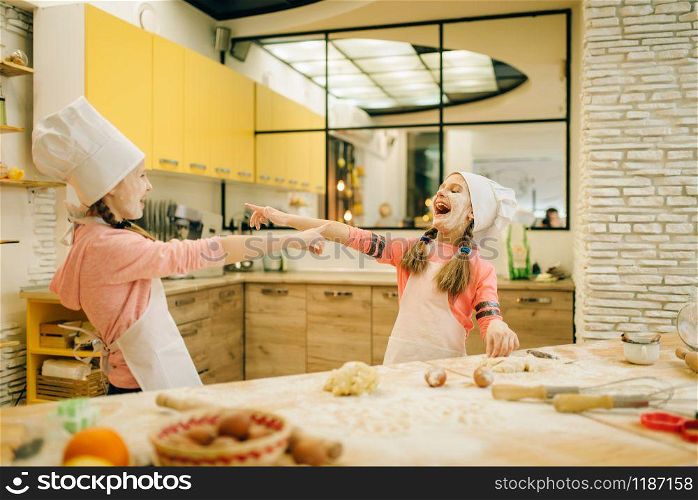 Two little girls chefs are laughing at each other, cookies preparation on the kitchen. Kids cooking pastry and having fun, children preparing cake