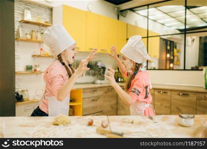 Two little girls chefs are plays and having fun, cookies preparation on the kitchen. Kids cooking pastry, children cooks preparing cake