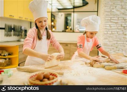 Two little girls chefs roll out the dough with rolling pins, cookies preparation on the kitchen. Kids cooking pastry and having fun, children cooks preparing cake