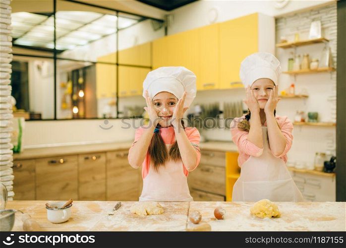 Two little girls chefs smears their faces with flour, cookies preparation on the kitchen. Kids cooking pastry and having fun, children preparing cake. Little girls chefs smears their faces with flour