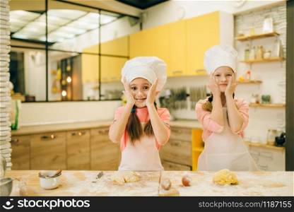 Two little girls chefs smears their faces with flour, cookies preparation on the kitchen. Kids cooking pastry and having fun, children preparing cake. Little girls chefs smears their faces with flour