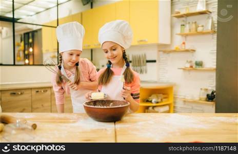 Two little girls cooks in caps adds butter to the bowl, cookies preparation on the kitchen. Kids cooking pastry, children chefs makes dough, child preparing cake. Little girls cooks in caps adds butter to the bowl