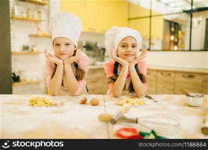 Two little girls cooks in caps are sitting at the table, cookies preparation on the kitchen. Kids cooking pastry, children chefs preparing cake
