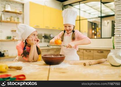 Two little girls cooks in caps rubs orange to the bowl, cookies preparation on the kitchen. Kids cooking pastry, children chefs preparing cake
