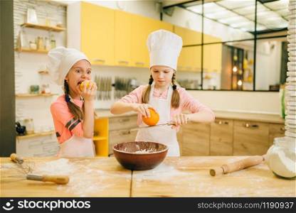Two little girls cooks in caps rubs orange to the bowl, cookies preparation on the kitchen. Kids cooking pastry, children chefs preparing cake. Little girls cooks in caps rubs orange to the bowl