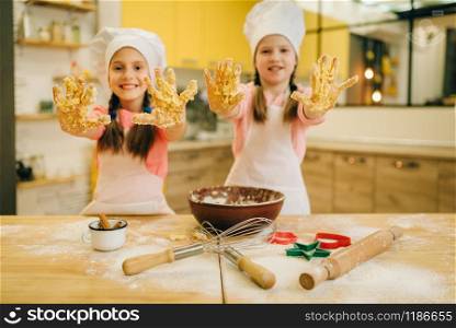 Two little girls cooks in caps shows hands covered in dough, cookies preparation on the kitchen. Kids cooking pastry, children chefs makes dough