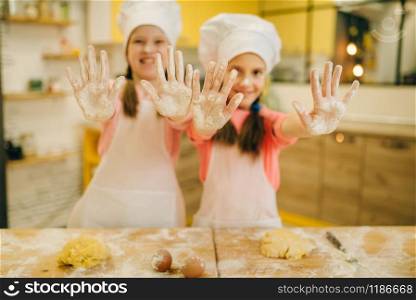 Two little girls cooks in caps shows hands covered in flour, cookies preparation on the kitchen. Kids cooking pastry, children chefs preparing cake