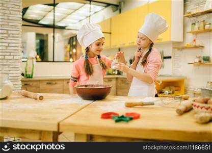 Two little girls cooks in caps tastes sweet vanilla powder, cookies preparation on the kitchen. Kids cooking pastry, children chefs preparing cake. Little girls cooks in caps tastes vanilla powder