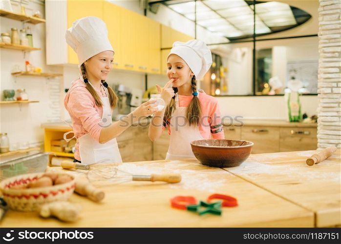 Two little girls cooks in caps tastes sweet vanilla powder, cookies preparation on the kitchen. Kids cooking pastry, children chefs preparing cake