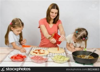 Two little girls enthusiastically watched as mum pours ketchup basis for pizza