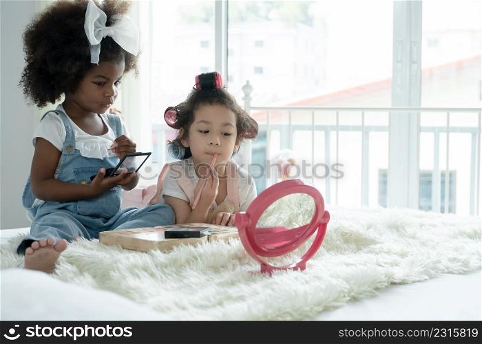 Two little girls friends, African and Caucasian, playing cosmetics together and use pink makeup brushes paint faces, lips, eyes, and cheeks with fun on bed at home. Ethnic diversity kids relationship