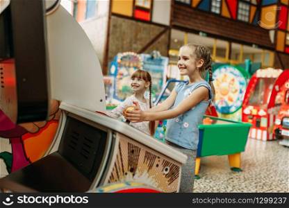 Two little girls plays on video game machine, children entertainment center. Excited childs having fun on playground indoors. Kids playing on motorcycles, amusement centre. Two little girls plays on video game machine