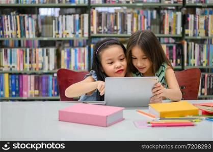 Two little happy cute girls playing on a tablet PC computing device in library at school. Education and self learning wireless technology concept. People lifestyles and friendship. Preschool children