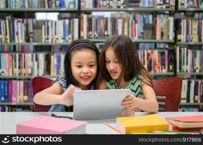 Two little happy cute girls playing on a tablet PC computing device in library at school. Education and self learning wireless technology concept. People lifestyles and friendship. Preschool children