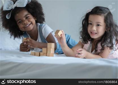 Two little kid girls, African and Asian children enjoy playing build wooden block toys together on bed at bedroom