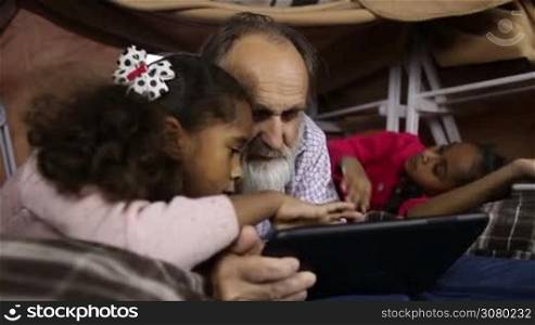 Two little mixed race girls explaining how to use digital tablet to handsome grandfather with beard. Serious children teaching grandpa to work on touchpad while lying down together in cubby house out of blanket and chairs. Dolly shot. Slo mo.
