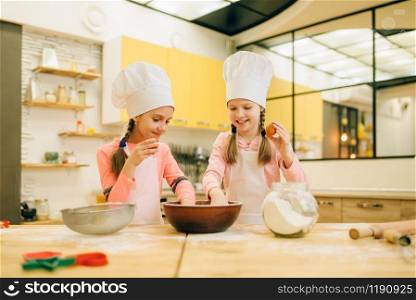 Two little sisters cooks in caps, cookies preparation on the kitchen. Kids cooking pastry, children chefs makes dough, child preparing cake