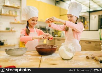 Two little sisters cooks in caps kneads eggs in a bowl, cookies preparation on the kitchen. Kids cooking pastry, children chefs makes dough, child preparing cake. Little sisters cooks in caps kneads eggs in a bow