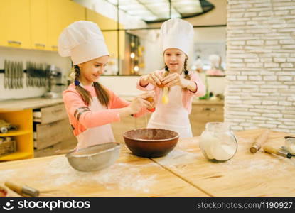 Two little sisters cooks in caps kneads eggs in a bowl, cookies preparation on the kitchen. Kids cooking pastry, children chefs makes dough, child preparing cake