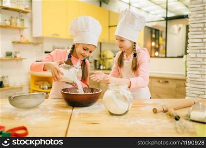 Two little sisters cooks in caps pours flour in a bowl, cookies preparation on the kitchen. Kids cooking pastry, children chefs makes dough, child preparing cake