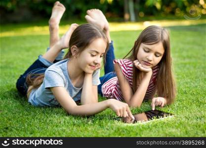 Two little sisters lying on grass outdoors and playing on digital tablet
