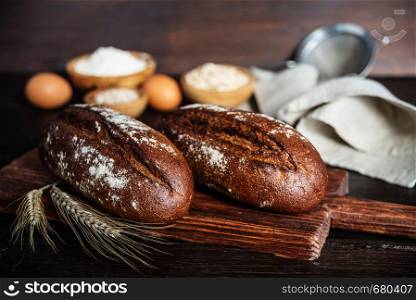 Two loaves of rye homemade bread, a gray linen napkin, flour and grain in wooden bowls, eggs and spikelets are on a dark wooden background
