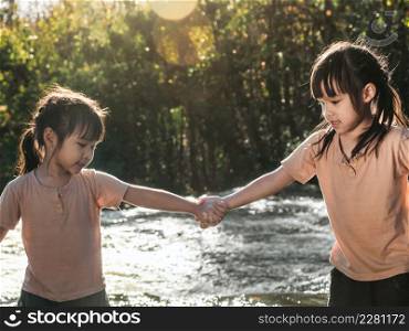 Two lovely girls holding hands happily playing in the stream. Asian sibling girls are playing in the waterfall in the forest. Active recreation with children on river in summer.