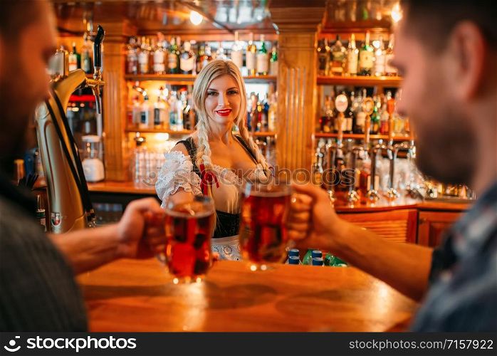 Two male friends clinking mugs with beer in pub, waitress at the counter on background, oktoberfest holidays. Men with glasses having fun in bar, barmaid in traditional retro style